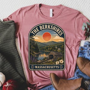 May include: A mauve t-shirt featuring a graphic of The Berkshires, Massachusetts, with a landscape of mountains, a lake, a covered bridge, and a sunset. The shirt is displayed with a dark green sweater, blue jeans, brown boots, and two small pumpkins.
