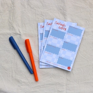 May include: A stack of three light blue and white checkered shopping list notepads with red text that reads Einkaufs LISTE and a blue pen and an orange pen placed to the left. The notepads feature sections for various grocery categories.