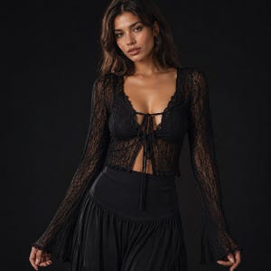 Puede incluir: Una blusa de encaje negro de manga larga con un escote en V profundo y cierre de lazo frontal. La prenda está confeccionada en encaje floral transparente y detallado, con mangas acampanadas de bordes festoneados y una falda negra.