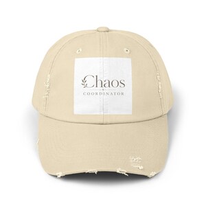 Könnte beinhalten: Eine beigefarbene, abgenutzte Baseballkappe mit einem weißen quadratischen Aufnäher auf der Vorderseite. Der Aufnäher zeigt den Text Chaos COORDINATOR in einer dunkelgrauen Serifenschrift, ergänzt durch eine kleine Blattgrafik links vom Wort Chaos.