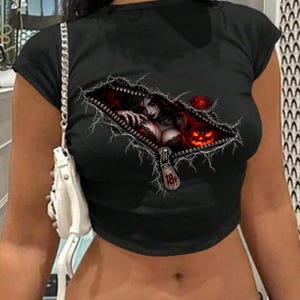 May include: A black short-sleeved crop top featuring a graphic print of a zipper opening to reveal a dark-haired figure and a glowing orange jack-o'-lantern. The zipper pull is marked with the text 18+.