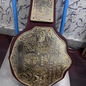 Puede incluir: Un cinturón de campeonato de lucha libre con una gran placa de metal dorado ornamentada con volutas intrincadas, un globo terráqueo y un emblema de corona. El texto dice WORLD HEAVYWEIGHT WRESTLING CHAMPION. Está montado sobre una correa de cuero granate.