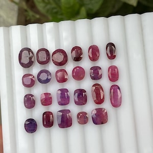 Op de afbeelding: Een verzameling van drieëntwintig gefacetteerde losse edelstenen in diverse tinten rood, roze en paars. Deze ovaal, kussen- en rond geslepen stenen zijn in een rasterpatroon gerangschikt op een wit geribbeld presentatieplateau.