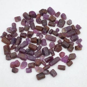 May include: A collection of numerous raw, unpolished gemstone crystals in various shades of deep red, purple, and brownish-pink. These small, irregularly shaped stones exhibit natural textures, varying levels of translucency, and faceted edges.