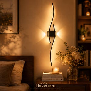 May include: A black wall-mounted light fitting featuring two slender, curved vertical bars that emit a warm, glowing light. The fitting is centred on a wall, casting a soft, symmetrical illumination pattern above and below the central square base.