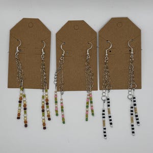 May include: Three pairs of dangle earrings displayed on brown cardboard tags. Each pair features silver-tone metal chains with dangling strands of small, multi-colored glass beads in earth tones, pink and green, and black, white, and gold patterns.