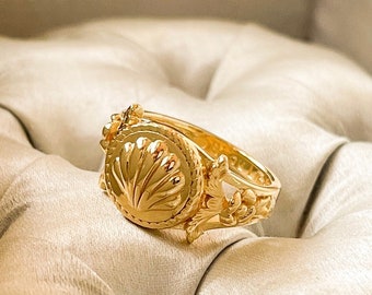 Könnte beinhalten: Ein goldener Ring mit einer runden Front, die ein erhabenes Muschelmotiv zeigt, umgeben von einem gedrehten Seilrand. Die Ringschiene ist auf beiden Seiten mit detaillierten floralen Schnitzereien verziert, die vom zentralen Motiv ausgehen.