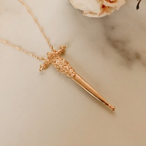 Puede incluir: Un collar de tono dorado con un colgante en forma de espada o daga delgada. El colgante presenta un mango texturizado con detalles intrincados tipo filigrana y una hoja lisa y puntiaguda. La cadena es fina y delicada en un tono dorado a juego.