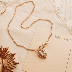 Op de afbeelding: Een goudkleurige ketting met een hartvormige hanger met een sierlijk ontwerp. De ketting ligt op een crèmekleurig oppervlak met een vintage ansichtkaart met de tekst "IMPRIME". Een vergrootglas is ook zichtbaar.