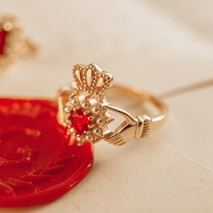May include: A gold ring with a red gemstone and white pearl accents. The ring is a Claddagh ring, a traditional Irish design featuring two hands clasping a heart.