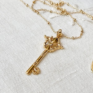 May include: A gold key necklace with a decorative, ornate key pendant. The chain is a delicate, beaded style.