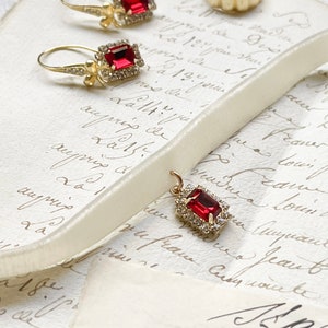 May include: A collection of gold jewelry featuring a pair of earrings, a ring, and a pendant. The earrings and pendant have a red rectangular stone surrounded by smaller clear stones. The ring is a simple gold band.