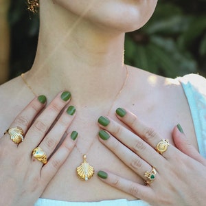 Op de afbeelding: Een gouden ketting met een schelp hanger en een parel accent. De ketting wordt gedragen door een persoon met groene nagellak. De persoon draagt ook verschillende gouden ringen met verschillende ontwerpen.