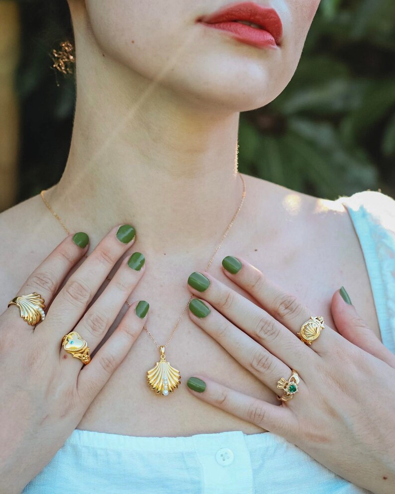 Op de afbeelding: Een gouden ketting met een schelp hanger en een parel accent. De ketting wordt gedragen door een persoon met groene nagellak. De persoon draagt ook verschillende gouden ringen met verschillende ontwerpen.