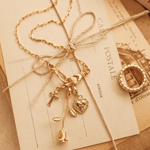 Puede incluir: Un collar dorado con un colgante de cruz, coraz&oacute;n y rosa, y un anillo a juego. El collar y el anillo est&aacute;n sobre una postal vintage con el texto "CAKE POSTALE" y "PARIS".