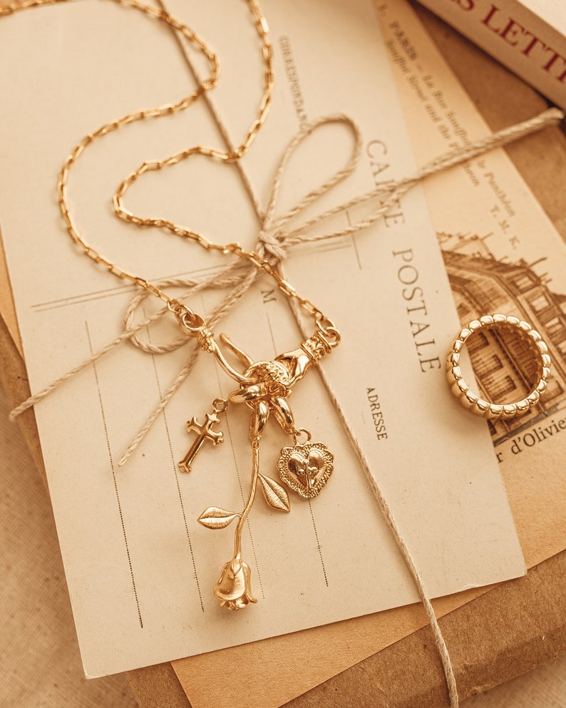 Puede incluir: Un collar dorado con un colgante de cruz, coraz&oacute;n y rosa, y un anillo a juego. El collar y el anillo est&aacute;n sobre una postal vintage con el texto "CAKE POSTALE" y "PARIS".