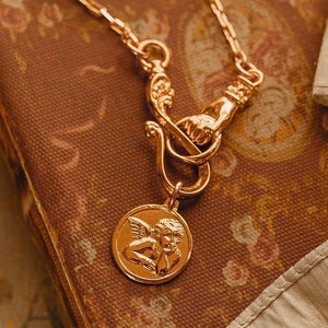 Op de afbeelding: Een gouden ketting met een gedetailleerde sluiting en een hanger met een cherubijn met vleugels. De hanger is rond en heeft een reliëfdesign.