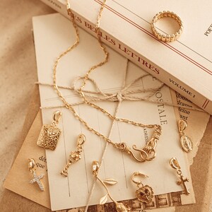 Puede incluir: Una colecci&oacute;n de joyas doradas, que incluye un anillo, collares y varios colgantes. Los colgantes presentan cruces, una rosa y s&iacute;mbolos religiosos. Los art&iacute;culos est&aacute;n dispuestos sobre papel vintage y un libro titulado "LES LETTRES".