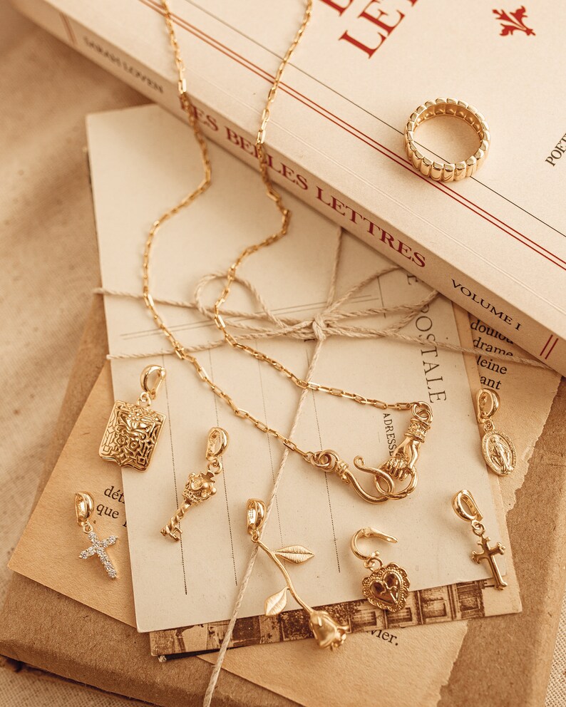 Puede incluir: Una colecci&oacute;n de joyas doradas, que incluye un anillo, collares y varios colgantes. Los colgantes presentan cruces, una rosa y s&iacute;mbolos religiosos. Los art&iacute;culos est&aacute;n dispuestos sobre papel vintage y un libro titulado "LES LETTRES".