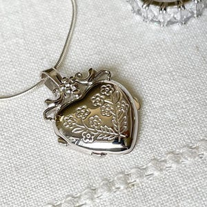May include: A silver heart-shaped locket necklace with a floral design. The locket has a detailed, ornate frame and is hanging on a delicate silver chain.
