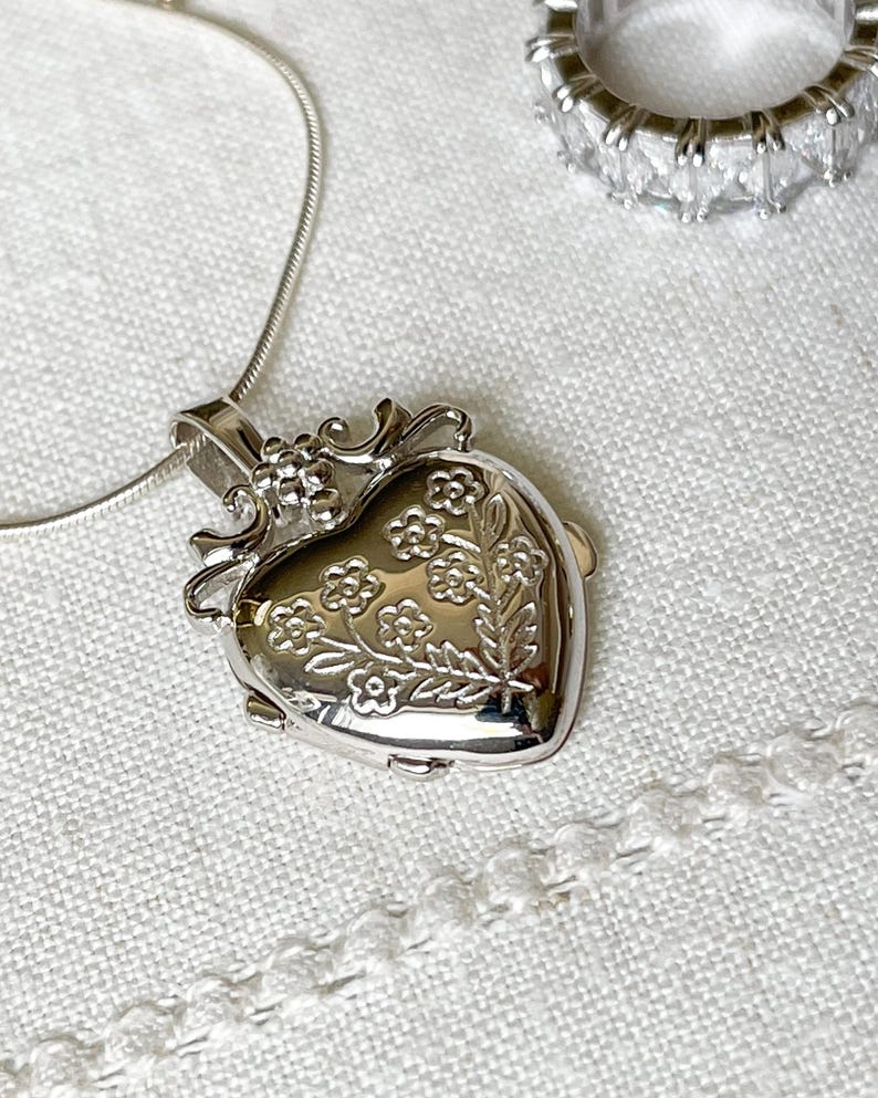 May include: A silver heart-shaped locket necklace with a floral design. The locket has a detailed, ornate frame and is hanging on a delicate silver chain.