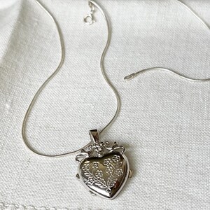 May include: A silver heart-shaped locket necklace with a delicate floral design. The chain is a snake chain style.