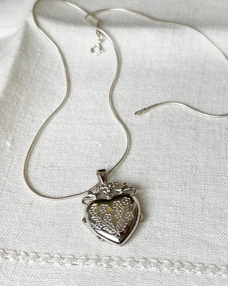 May include: A silver heart-shaped locket necklace with a delicate floral design. The chain is a snake chain style.