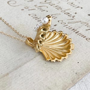 Op de afbeelding: Een gouden ketting met een schelp hanger die opengaat om een ​​kleine parel te onthullen. De ketting ligt op een wit oppervlak met schrift.