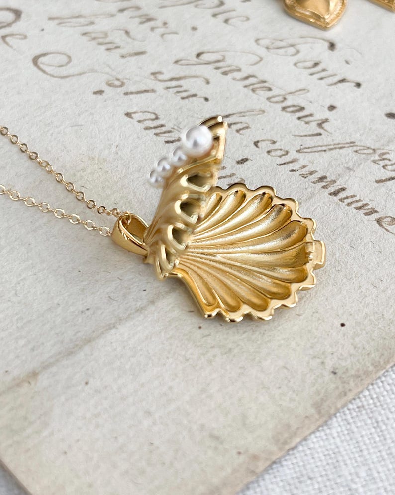 Op de afbeelding: Een gouden ketting met een schelp hanger die opengaat om een ​​kleine parel te onthullen. De ketting ligt op een wit oppervlak met schrift.