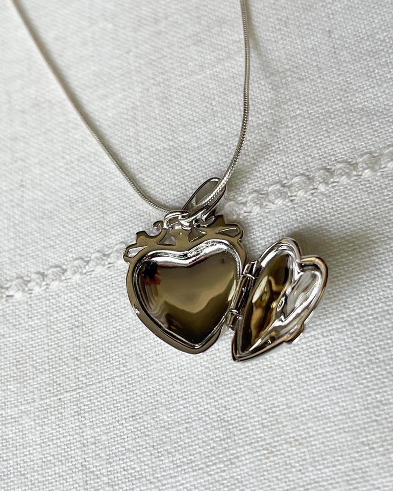 May include: A silver heart-shaped locket necklace with a hinged lid open, revealing the empty interior. The chain is a delicate silver snake chain.