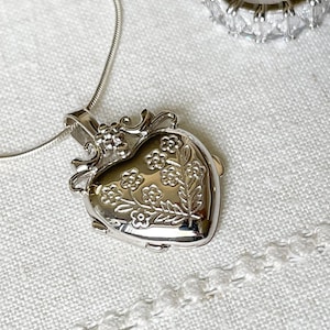 May include: A silver heart-shaped locket necklace with a floral design. The locket has a detailed, ornate frame and is hanging on a delicate silver chain.