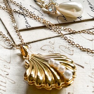 Op de afbeelding: Een gouden ketting met een schelp-hanger met drie witte parels. De hanger is open en heeft een gedetailleerd ontwerp. De ketting ligt op een vintage-achtig oppervlak.