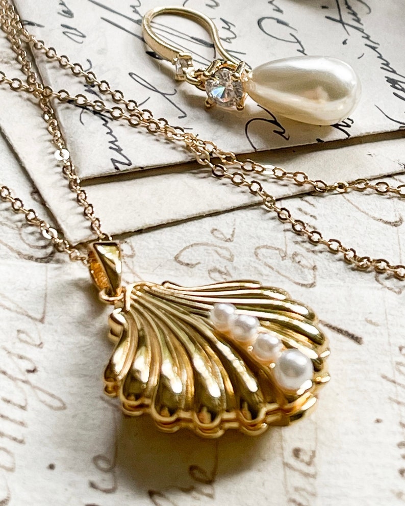 Op de afbeelding: Een gouden ketting met een schelp-hanger met drie witte parels. De hanger is open en heeft een gedetailleerd ontwerp. De ketting ligt op een vintage-achtig oppervlak.