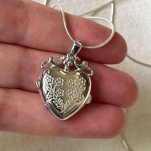 May include: A silver heart-shaped locket necklace with a floral design. The locket has a hinged opening and a delicate chain.