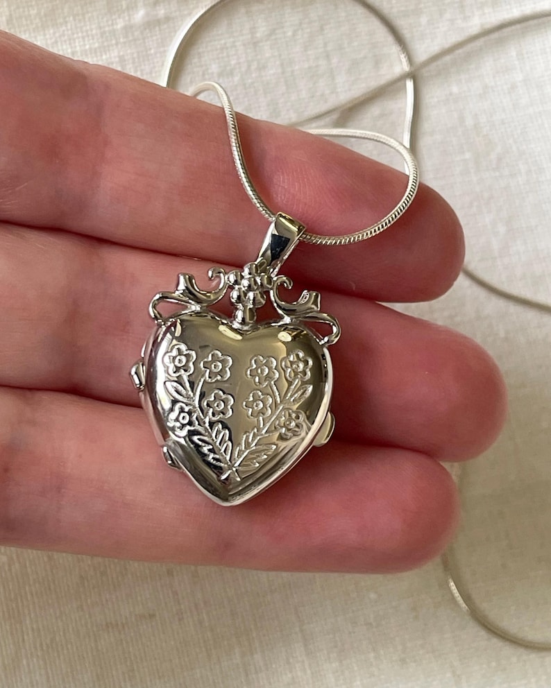 May include: A silver heart-shaped locket necklace with a floral design. The locket has a hinged opening and a delicate chain.