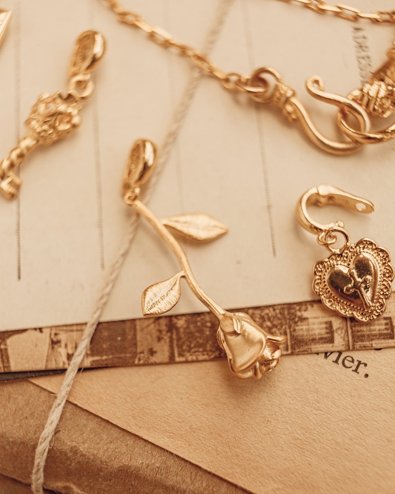 Puede incluir: Primer plano de joyas doradas. Se ven un colgante de rosa, un pendiente en forma de coraz&oacute;n y un dije en forma de llave. Las joyas est&aacute;n colocadas sobre un fondo de papel y cuerda vintage, creando una est&eacute;tica cl&aacute;sica.