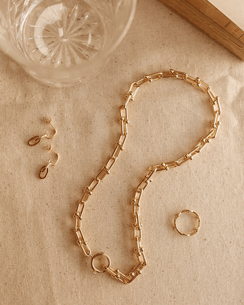 K&ouml;nnte beinhalten: Ein goldfarbenes Schmuckset mit einer Kette, einem Ring und einem Paar Ohrringen. Die Halskette hat einen gro&szlig;en Verschluss und ein Kettenglied-Design. Der Ring hat ein verdrehtes Design, und die Ohrringe sind klein und zart. Der Schmuck wird auf einer beigen Oberfl&auml;che pr&auml;sentiert.