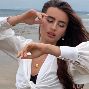 Op de afbeelding: Een vrouw draagt een wit blouse met een geknoopte voorkant en een gouden schelpketting. Ze staat op een strand met de zee op de achtergrond.