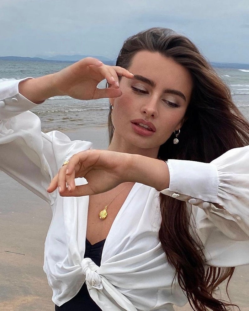 Op de afbeelding: Een vrouw draagt een wit blouse met een geknoopte voorkant en een gouden schelpketting. Ze staat op een strand met de zee op de achtergrond.