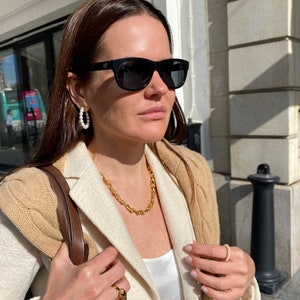 K&ouml;nnte beinhalten: Eine Frau mit schwarzer Sonnenbrille, Perlenohrringen und einer goldenen Halskette. Sie tr&auml;gt au&szlig;erdem einen beigefarbenen und wei&szlig;en Blazer und einen goldenen Ring. Eine braune Ledertasche h&auml;ngt &uuml;ber ihrer Schulter.