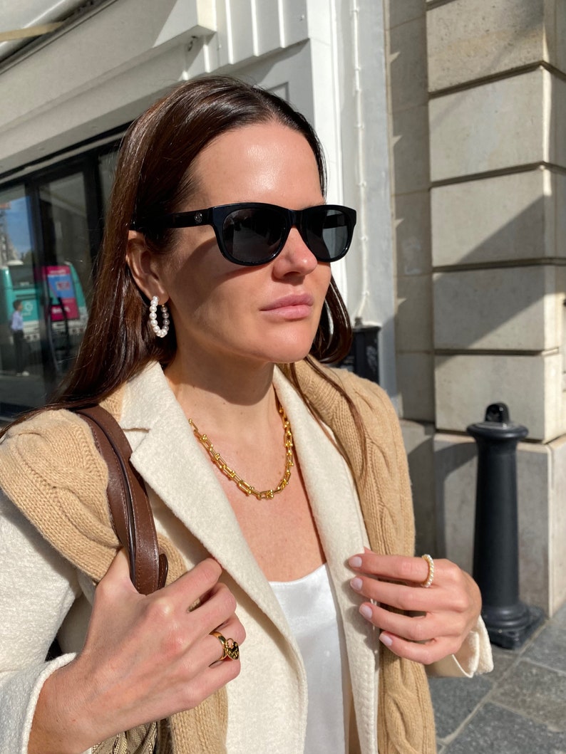 K&ouml;nnte beinhalten: Eine Frau mit schwarzer Sonnenbrille, Perlenohrringen und einer goldenen Halskette. Sie tr&auml;gt au&szlig;erdem einen beigefarbenen und wei&szlig;en Blazer und einen goldenen Ring. Eine braune Ledertasche h&auml;ngt &uuml;ber ihrer Schulter.