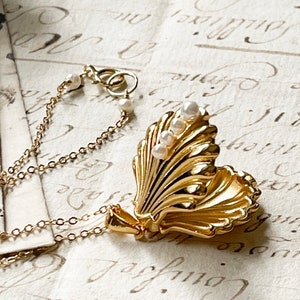 Op de afbeelding: Een gouden schelp-hangerketting met een ketting en parel accenten. De hanger kan worden geopend om een klein compartiment aan de binnenkant te onthullen.