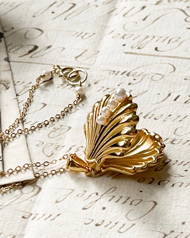 Op de afbeelding: Een gouden schelp-hangerketting met een ketting en parel accenten. De hanger kan worden geopend om een klein compartiment aan de binnenkant te onthullen.