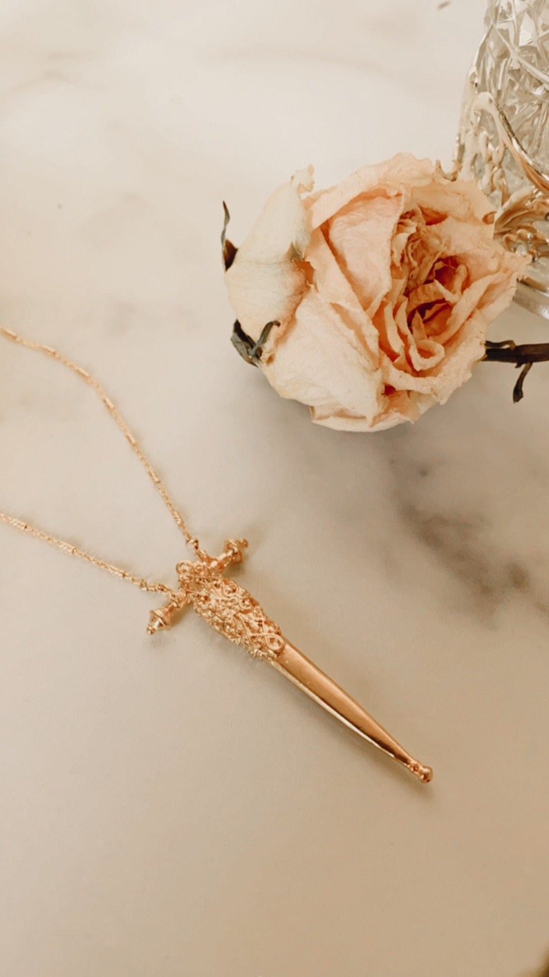 Puede incluir: Un collar de tono dorado con un colgante en forma de espada o daga delgada. El colgante presenta un mango texturizado con detalles intrincados tipo filigrana y una hoja lisa y puntiaguda. La cadena es fina y delicada en un tono dorado a juego.
