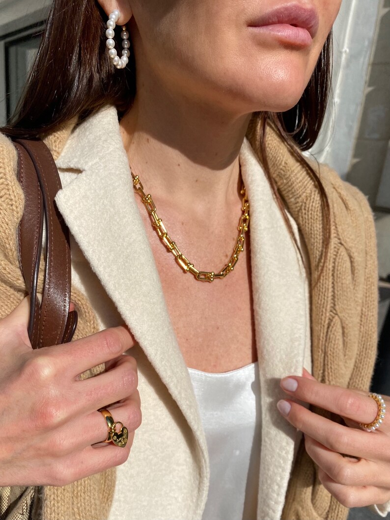 K&ouml;nnte beinhalten: Eine Person tr&auml;gt Goldschmuck, darunter eine Kette, Creolen und einen Ring. Sie tr&auml;gt auch einen beigefarbenen Zopfstrickpullover und einen wei&szlig;en Blazer. Der Schmuck scheint goldfarben zu sein und die Ohrringe sind aus Perlen.