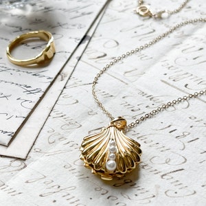 Op de afbeelding: Een gouden ketting met een schelp-hanger. Het medaillon is open en bevat drie witte parels. De ketting ligt op een witte achtergrond.