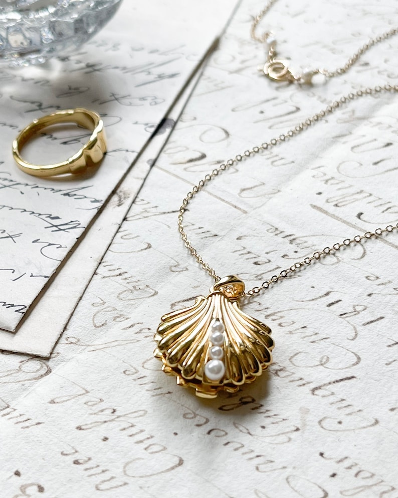 Op de afbeelding: Een gouden ketting met een schelp-hanger. Het medaillon is open en bevat drie witte parels. De ketting ligt op een witte achtergrond.