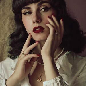 Op de afbeelding: Een vrouw met donker haar en rode lippenstift draagt een wit blouse met een parelketting en een gouden schelp hanger. Ze draagt gouden ringen aan haar vingers en heeft een neuspiercing.
