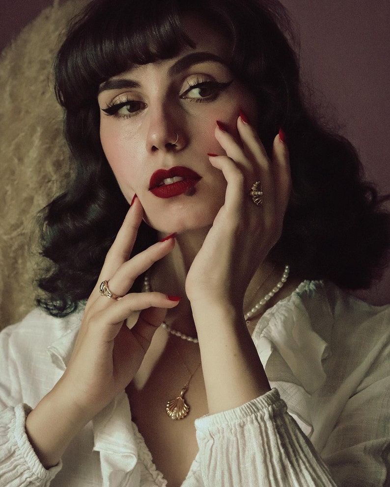 Op de afbeelding: Een vrouw met donker haar en rode lippenstift draagt een wit blouse met een parelketting en een gouden schelp hanger. Ze draagt gouden ringen aan haar vingers en heeft een neuspiercing.