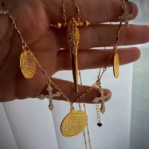 Puede incluir: Un collar de tono dorado que presenta un colgante central en forma de espada con detalles de filigrana, acentuado por m&uacute;ltiples dijes redondos estilo moneda y peque&ntilde;os colgantes de cruz incrustados con piedras claras brillantes.